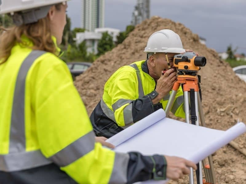 Surveyor using total station equipment while reviewing site plans at a commercial property site for ALTA survey due diligence and risk review.